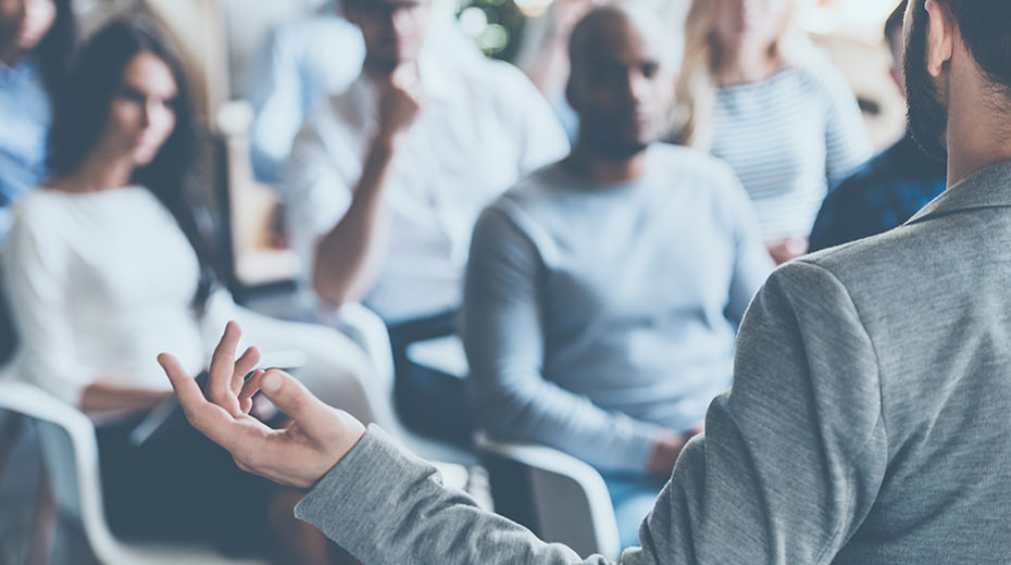 The back of a man in a suit facing a crowd of people as he speaks. This symbolizes how sharing the expertise of your in-house experts can create brand awareness.